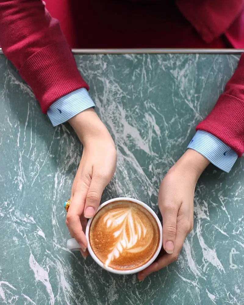 در کافه ث، دستانی یک فنجان قهوه لاته آرت جذاب را روی میز مرمر سبز رنگ در فضایی دلنشین و آرامش‌بخش گرفته‌اند.