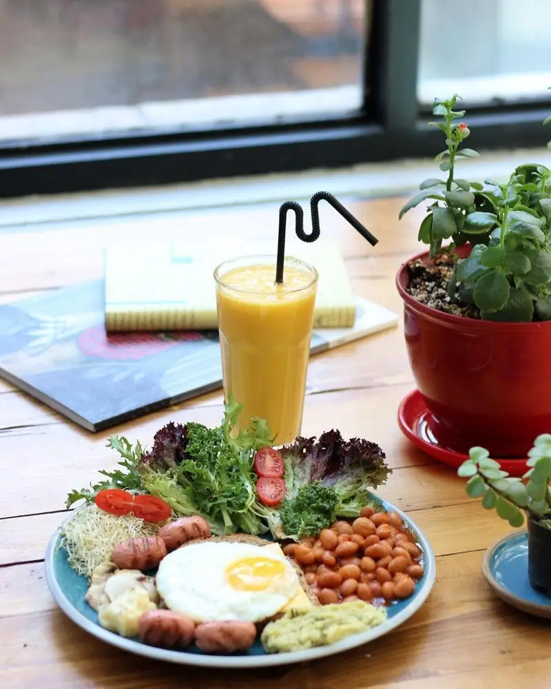 یک صبحانه مفصل و سالم کافه ث: نیمرو، سوسیس، لوبیا پخته، سالاد تازه و آب پرتقال کنار پنجره با نور طبیعی.