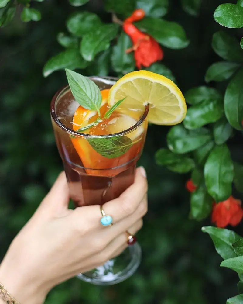 نوشیدنی گوارای کافه ث: چای سرد لیمو و پرتقال با برگ‌های تازه نعناع، سرو شده در فضای باز و دلنشین باغی سرسبز با گلهای انار.