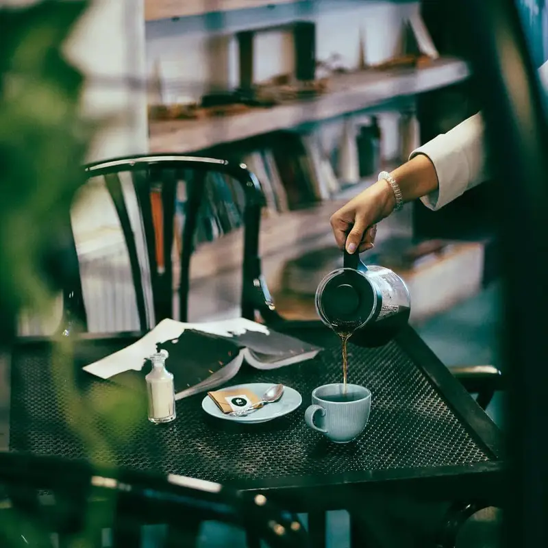 لحظه‌ای دلپذیر در کافه ث: دست در حال ریختن قهوه تازه در فنجان، همراه با کتاب و وسایل نوشیدنی روی میز با بافت خاص، فضایی دنج و آرام.