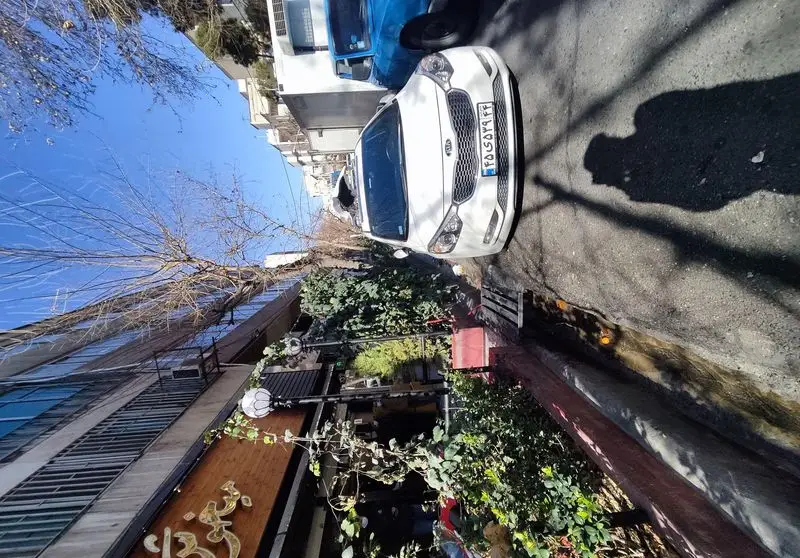 تصویری از نمای بیرونی فست فود پیتزا میخوش در خیابان، با خودروهای پارک شده، آسمان آبی و درختان در روز آفتابی.