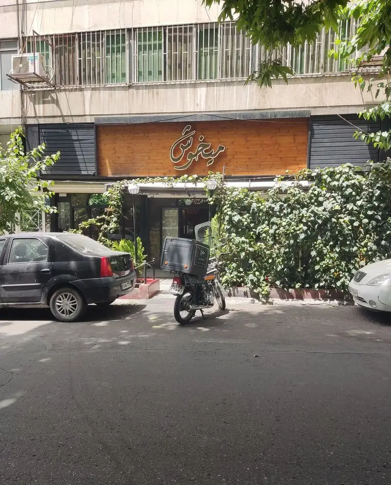 تصویری از نمای بیرونی فست فود پیتزا میخوش در یک روز آفتابی، با تابلوی چوبی زیبا و گیاهان سبز. موتورسیکلت دلیوری نیز در تصویر دیده می‌شود.