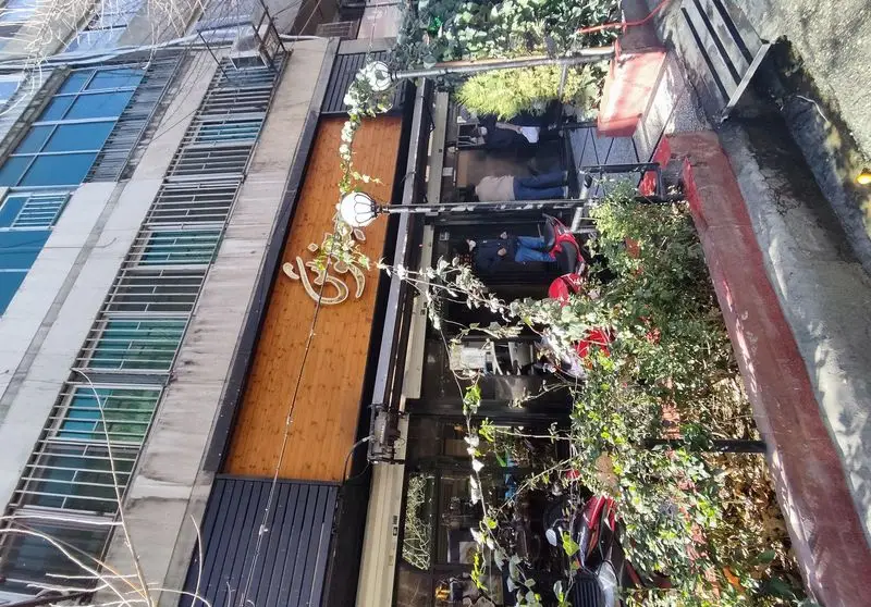 نمای بیرونی فست فود پیتزا میخوش با تابلوی جذاب، تزئینات گیاهی، و مشتریان در فضای باز. ساختمانی با پنجره‌های آبی و چوب.