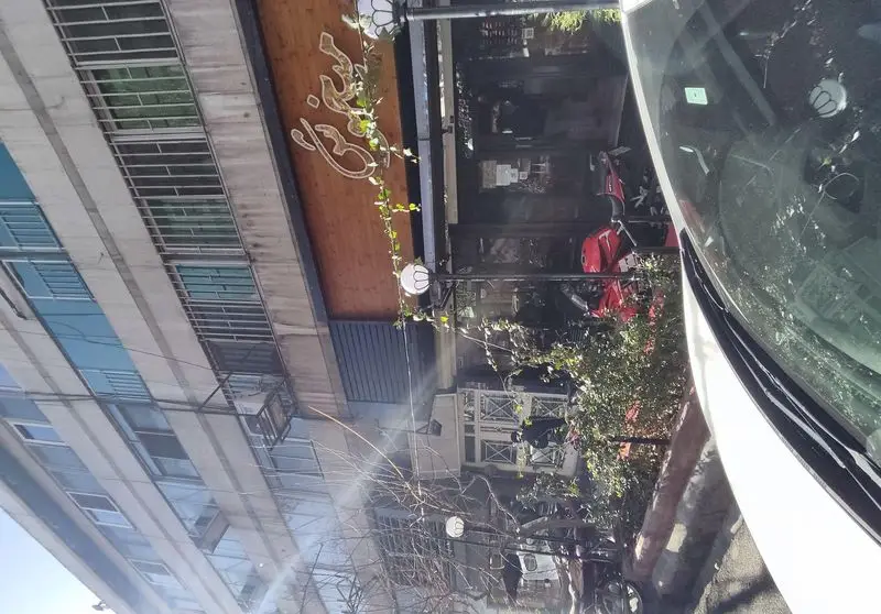 نمای بیرونی فست فود پیتزا میخوش با تابلوی چوبی، گیاهان آویز، چراغ‌های خیابانی و یک اسکوتر قرمز پارک شده در جلوی مغازه