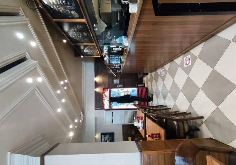 فضای داخلی مدرن فست فود پیتزا میخوش با پیشخوان شیک، منوی واضح، صندلی‌ها و مشتری کنار یخچال نوشیدنی.