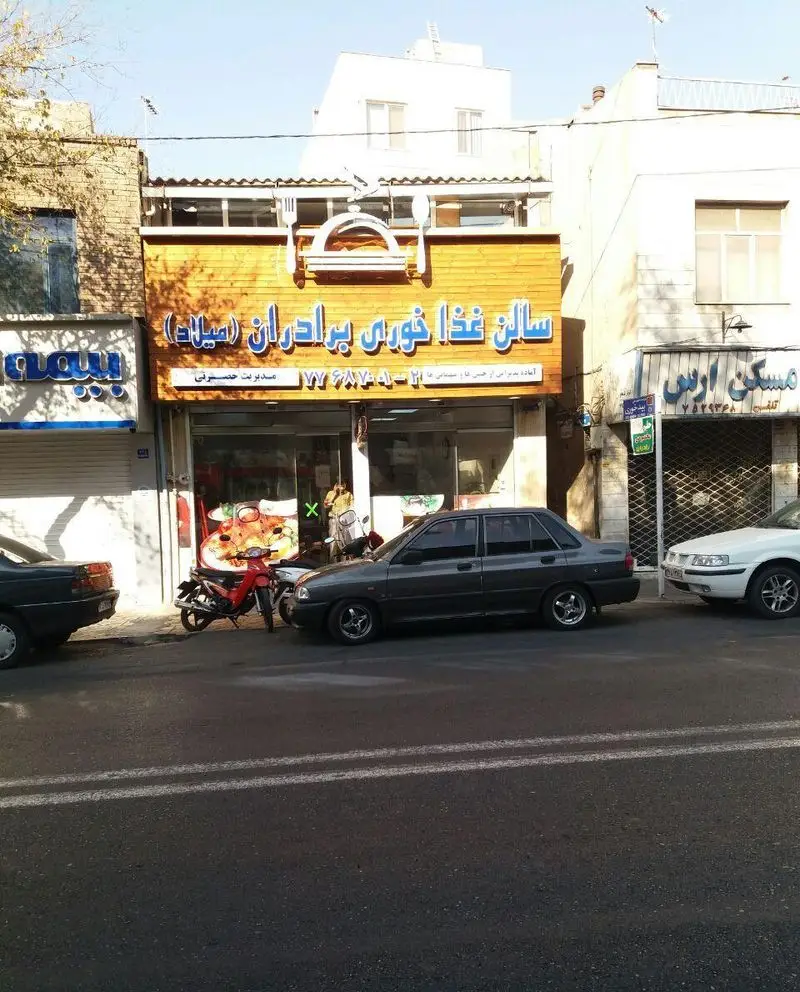 تصویری از نمای بیرونی سالن غذاخوری برادران میلاد۲ در تهران، با تابلوی چوبی و آبی، ارائه دهنده غذای ایرانی و پذیرایی مجالس.