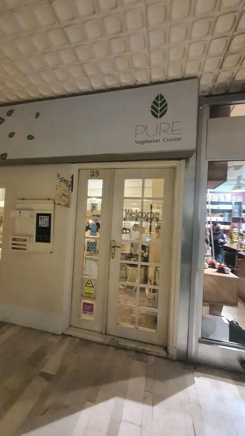 نمای بیرونی رستوران گیاهی پیور، با تابلوی روشن "PURE Vegetarian Cuisine" و لوگوی برگ سبز. درب ورودی شماره 35 رستوران با کدهای QR برای دسترسی به منو قابل مشاهده است.