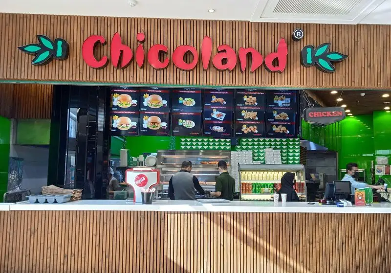 تصویری از رستوران چیکولند در فودکورت سانا، شامل منوی دیجیتال برگر و مرغ سوخاری، کارکنان پشت پیشخوان سفارش در محیطی مدرن.