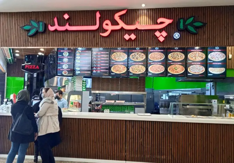 نمای داخلی رستوران فست فود چیکولند فودکورت سانا با تابلو نئون، منوی دیجیتال پیتزا و مشتریان در کانتر سفارش غذا.