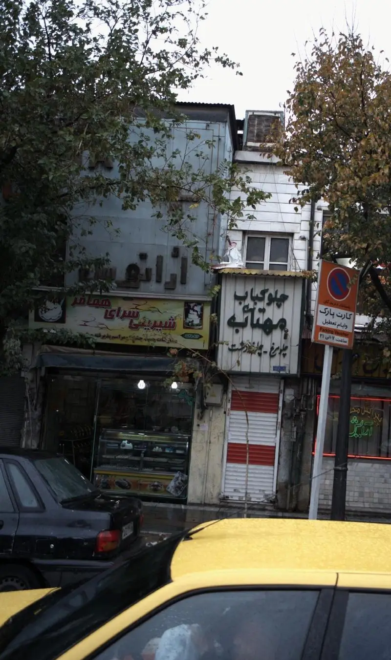 تصویر خیابان با شیرینی سرای همافر، ویترین شلوغ شیرینی، چلوکباب مهتابه کنار آن، درختان و تاکسی زرد در پیش‌زمینه.