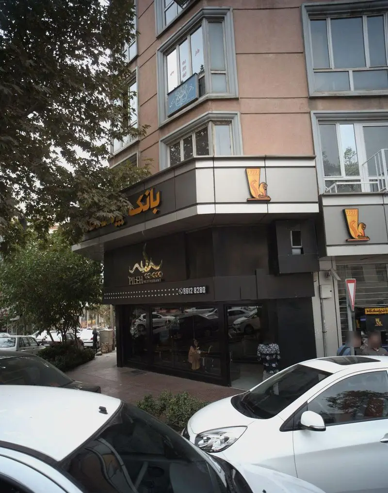 نمای بیرونی فست فود پیله (Pileh Fast Food) در یک روز آفتابی با ماشین‌های پارک شده و درختان. تابلوی رستوران و شماره 86128280.