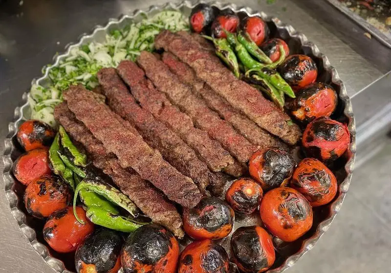 تصویری جذاب از کباب کوبیده داغ و لذیذ، همراه با گوجه‌های آبدار و فلفل‌های سبز کبابی، روی سینی فلزی بزرگ، از کباب و جگر سرای عمو هوشنگ.