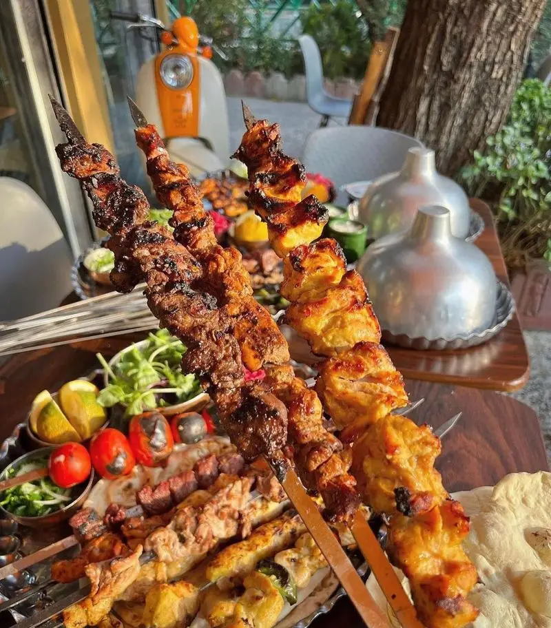 کباب و جگر سرای عمو هوشنگ: کباب‌های لذیذ کوبیده و جوجه زعفرانی به همراه گوجه کبابی و نان داغ، میز شلوغ در فضای باز.