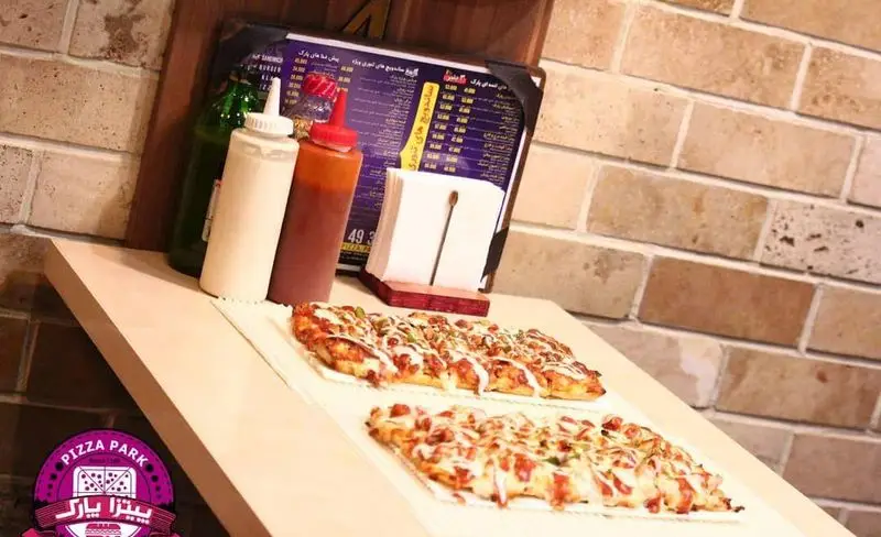 پیتزا پارک: تصویر دو پیتزای مستطیلی داغ و خوشمزه روی میز، با سس مایونز و کچاپ، منو رستوران و دیوار آجری فست فود.