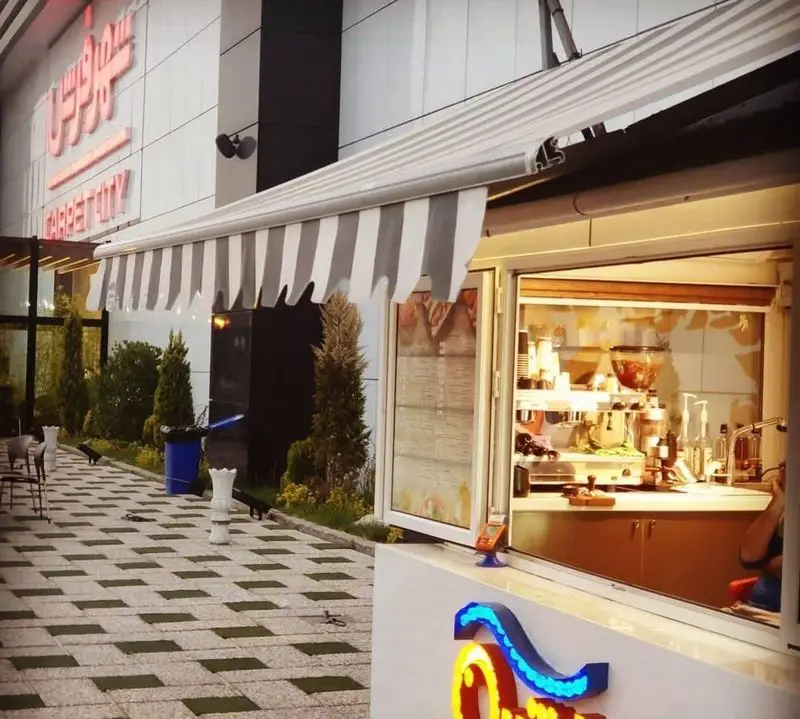 کافه رستوران آرشیو: نمای جذاب فضای باز با سایبان مدرن و راه‌راه، میز و صندلی و ویترین آماده سرویس‌دهی.