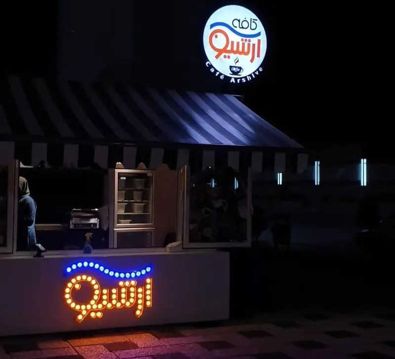 تصویری جذاب از کافه رستوران آرشیو در شب، با تابلوی بزرگ نورانی "کافه آرشیو" و تابلوی نئون "آرشیو" روی پیشخوان، فضایی دلنشین برای مشتریان.