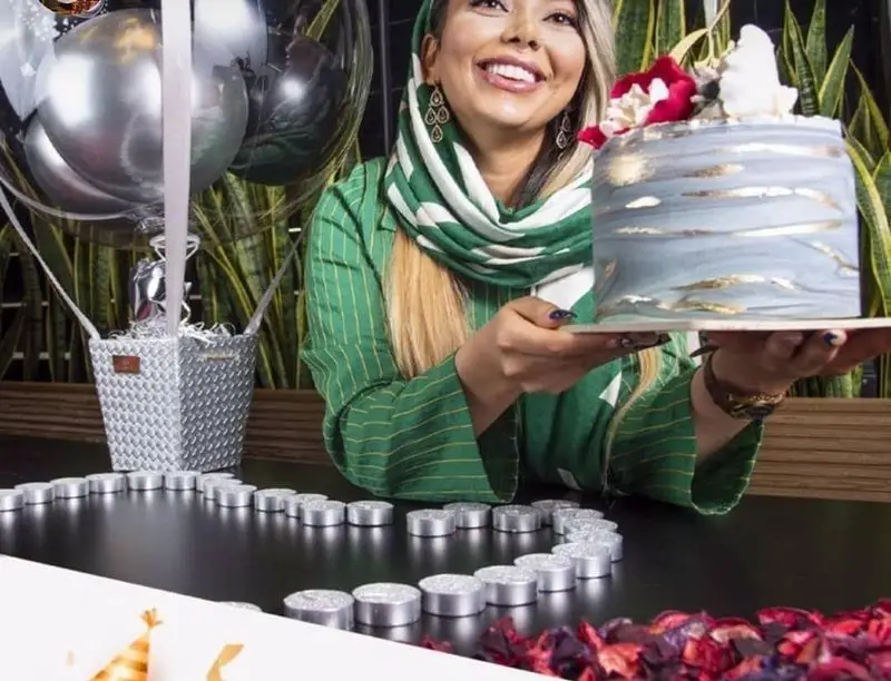 تصویری شاد از مشتری در فودهال نارسیس، کیک تولد یا مناسبتی با تزیینات خاص و آبی-طلایی را در دست دارد. محیط با شمع و بالن تزئین شده.