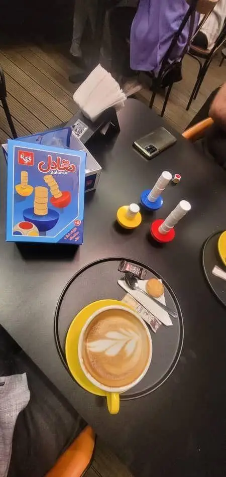 بازی رومیزی تعادل و فنجان لاته آرت زیبا روی میز در فودهال نارسیس، محیطی دوستانه و سرگرم‌کننده برای دورهمی‌های کافه را به تصویر می‌کشد.