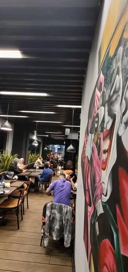 نمایی از فضای دنج و شیک فودهال نارسیس، مشتریان در حال لذت بردن از غذا و نوشیدنی با دکوراسیون مدرن و نقاشی جذاب جوکر.