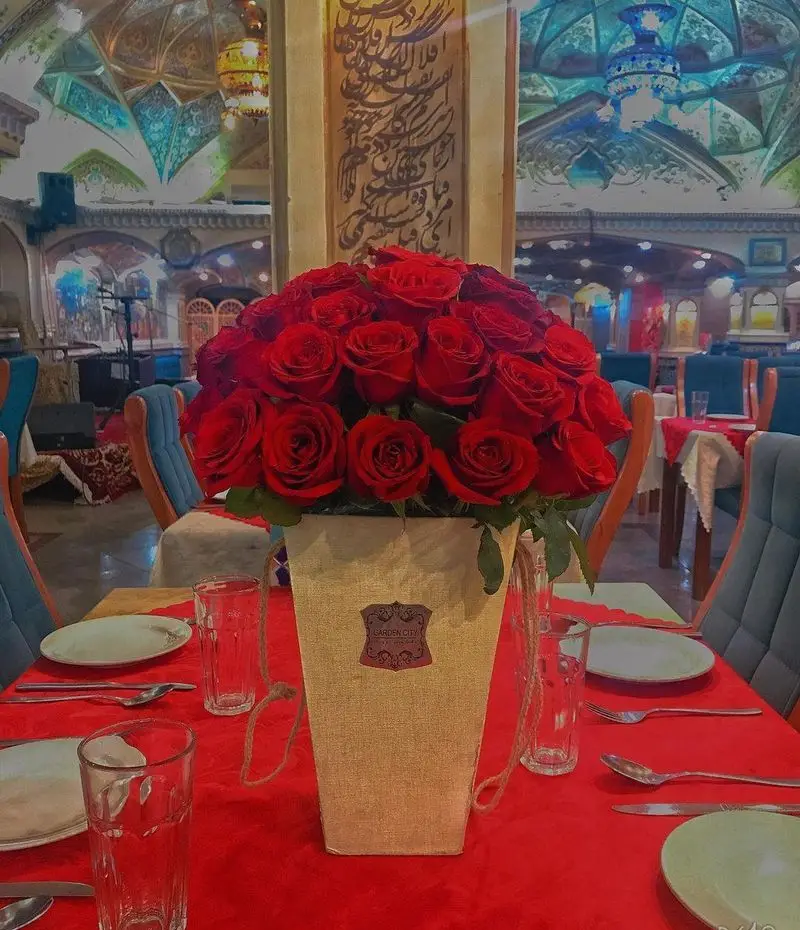 نمایی از دکوراسیون داخلی رستوران دف با میزهای چیدمان شده، گلدان گل رز قرمز، و معماری سنتی ایرانی برای تجربه‌ای دلنشین.