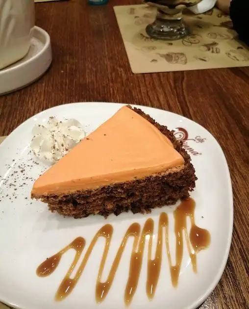 برش کیک دسر پرتقالی با پایه شکلاتی و سس کارامل، همراه با خامه، سرو شده در کافه ویونا؛ لذت طعم های جدید.