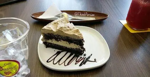 برش کیک شکلاتی با خامه و سس شکلات در کافه ویونا، روی میز چوبی. لیوان آب یخ با برچسب "کافه ویونا" و یک نوشیدنی قرمز نیز در تصویر هستند.