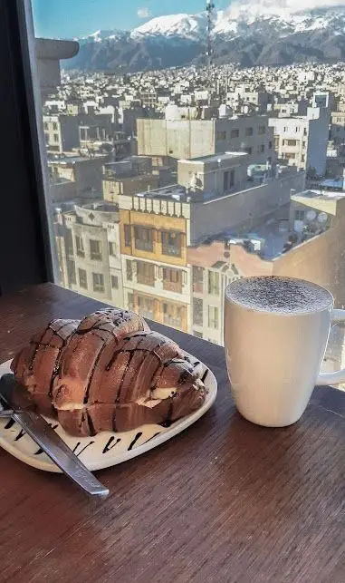 لذت صبحانه یا عصرانه با کروسان شکلاتی و قهوه در کافه ویونا، همراه با منظره بی‌نظیر شهر و کوهستان برفی.