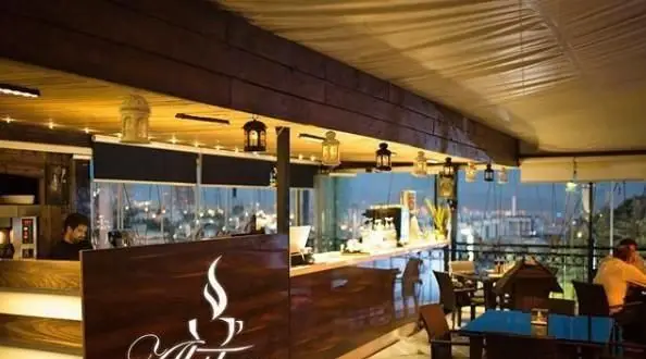 فضای داخلی دنج کافی شاپ آبشار با دکوراسیون جذاب، منظره زیبای شهر در شب و میزهای چوبی شیک برای لذت بردن از قهوه.