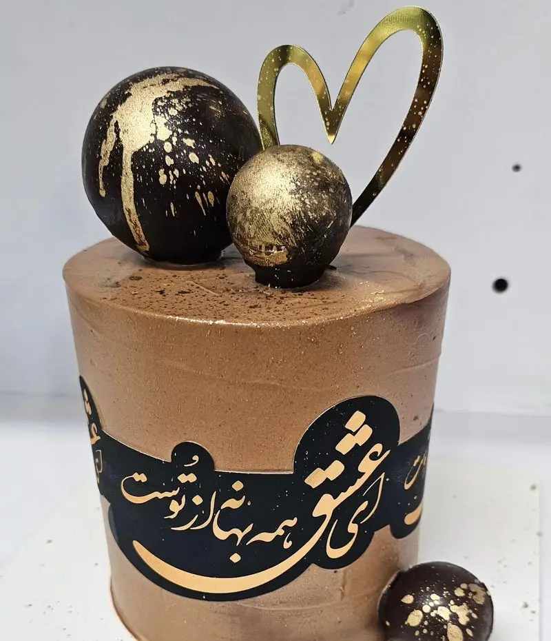 کیک سفارشی شکلاتی قهوه‌ای شیرینی خاتون، با تزیین گوی‌های طلایی و تاپر قلب، و خطاطی "عشق یعنی همه بها از توست".
