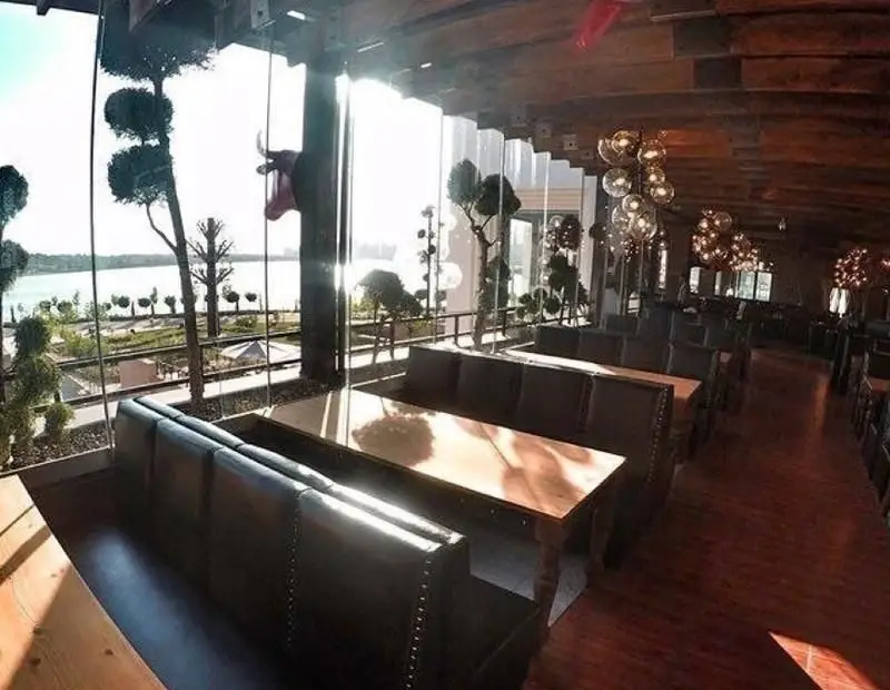 نمای داخلی رستوران ایرانی ته دیگ با میزهای چوبی و صندلی‌های راحتی، چشم‌انداز زیبای دریاچه و دکوراسیون مدرن و شیک برای تجربه‌ای لذت‌بخش.