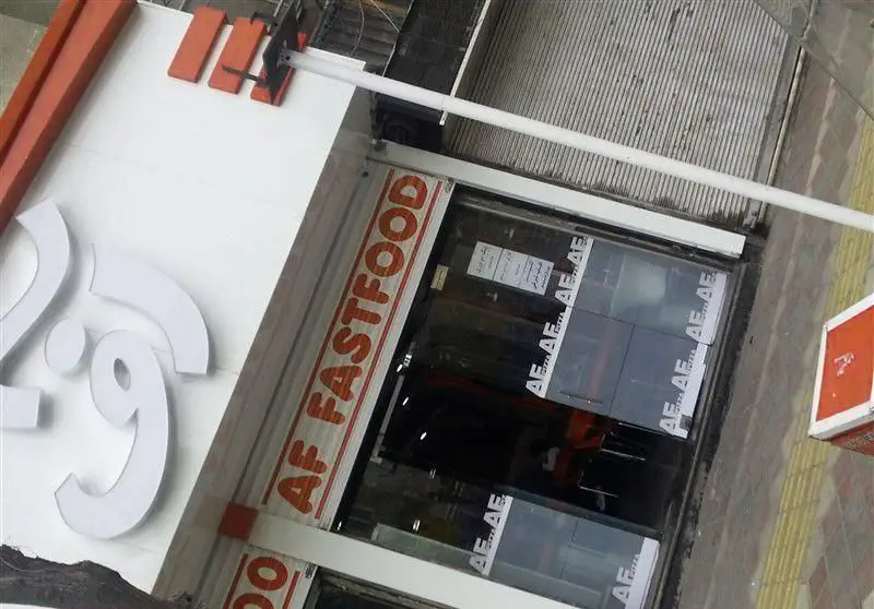 تصویری از نمای بیرونی فست فود اف، با تابلوی سفید بزرگ و نام 'AF FASTFOOD' بالای درب ورودی، و جزئیات تکراری 'AF' روی شیشه های ورودی.