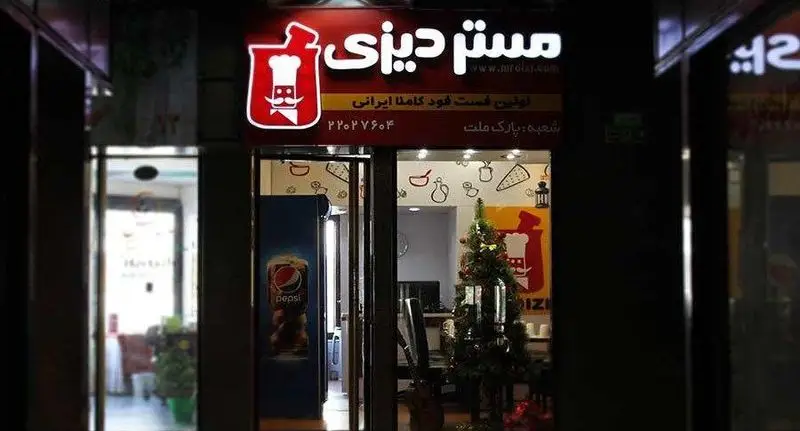 تصویری از نمای شبانه ورودی رستوران مستر دیزی شعبه پارک ملت، با تابلوی قرمز رنگ و لوگوی سرآشپز. این مکان اولین فست فود ایرانی است.