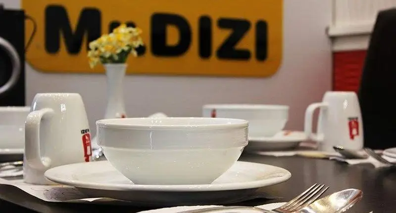 چیدمان شیک میز در رستوران مستر دیزی شعبه پارک ملت، با ظروف غذاخوری تمیز و براق و لوگوی بزرگ مستر دیزی در پس زمینه.