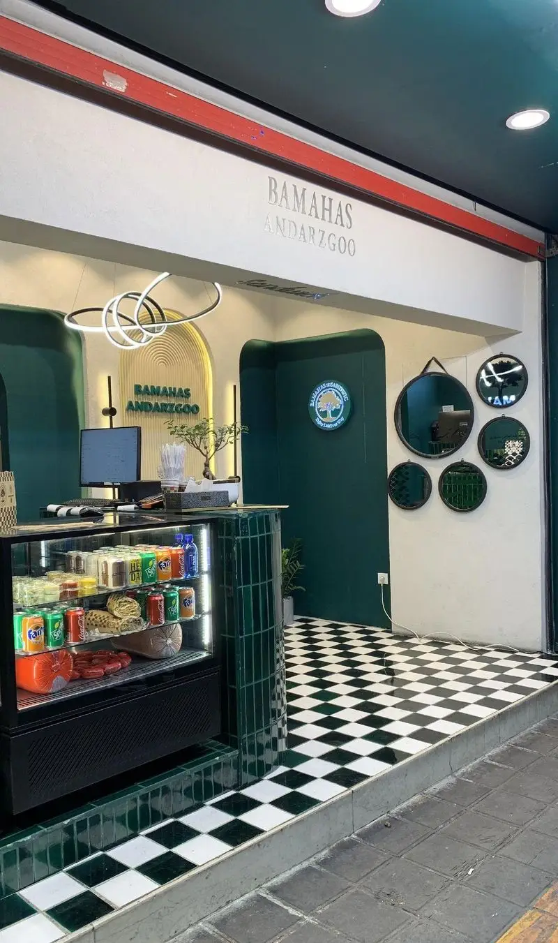 تصویری از نمای داخلی و ورودی باماهاس اندرزگو، فست فود و کافه ساندویچ مدرن در تهران با کفپوش شطرنجی، پیشخوان و یخچال نمایش.