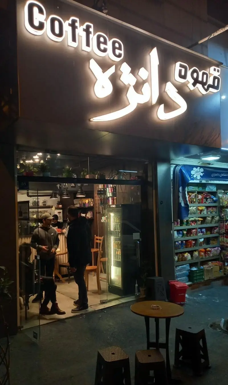 نمای شب کافه دانژه با تابلوی نئونی "Coffee" و "قهوه دانژه". مشتریان در فضای داخلی و خارجی مشغول نوشیدن قهوه. محیط دنج و دعوت‌کننده.