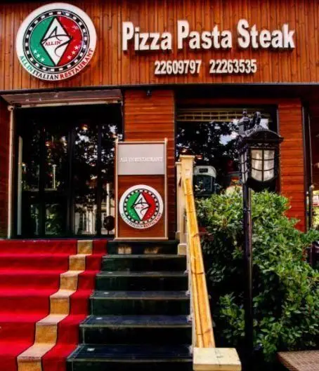تصویر بیرونی رستوران ایتالیایی آلین شامل تابلوی "پیتزا، پاستا، استیک" با شماره تلفن‌های 22609797 و 22263535 و ورودی شیک با فرش قرمز.