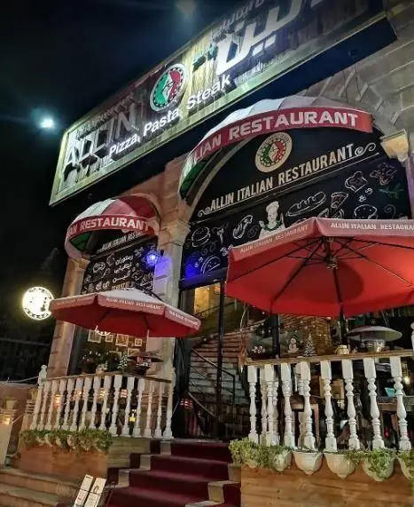 ورودی شبانه و نورانی رستوران ایتالیایی آلین با چترهای قرمز، تابلوهای پیتزا، پاستا و استیک، فضایی دلنشین برای مشتریان.