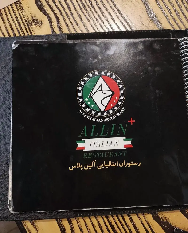 کاور منوی رستوران ایتالیایی آلین پلاس، با طراحی شیک و چشم‌نواز شامل لوگوی پرچم ایتالیا و نام برند، روی میز چوبی رستوران.