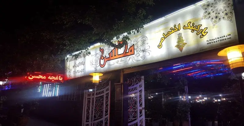 نمای شبانه از ورودی باغچه محسن با تابلوی روشن، پارکینگ اختصاصی و نورپردازی زیبا، مکانی دنج برای رستوران و کافه.