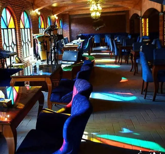 تصویری از فضای داخلی دلپذیر باغچه محسن، یک رستوران سنتی با پنجره‌های رنگی، نورپردازی شاداب و مبلمان آبی راحت.