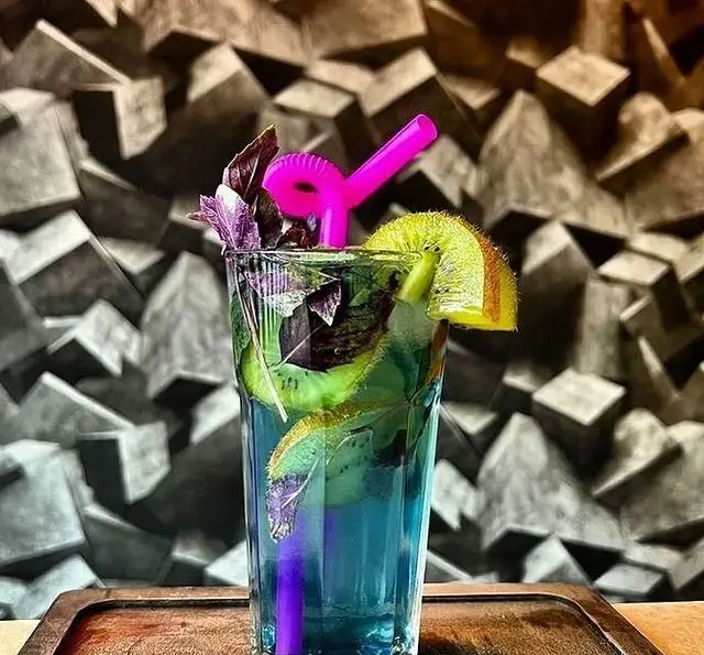 تصویری جذاب از نوشیدنی خنک و آبی‌رنگ باغچه محسن، تزئین شده با برش‌های تازه کیوی، برگ‌های معطر ریحان بنفش و نی فانتزی صورتی. مناسب برای روزهای گرم تابستان.