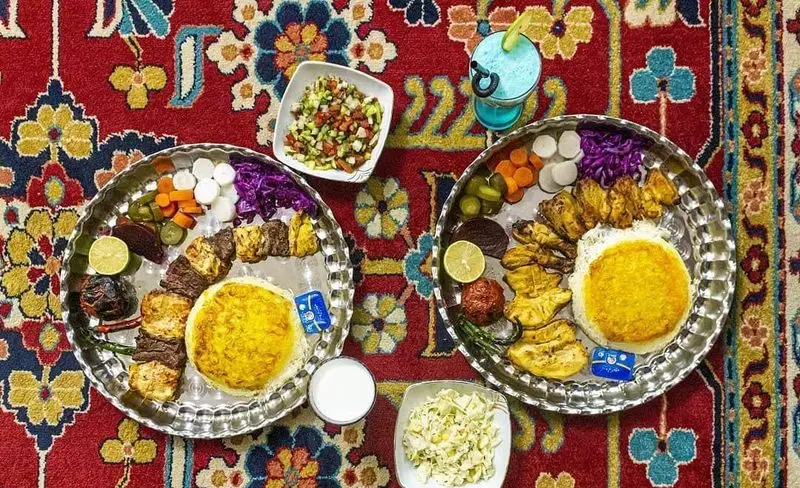پذیرایی سنتی گل زعفرون: دو سینی کباب لذیذ (جوجه و کوبیده)، برنج زعفرانی، ته دیگ، ترشی و سالاد روی فرش اصیل ایرانی.
