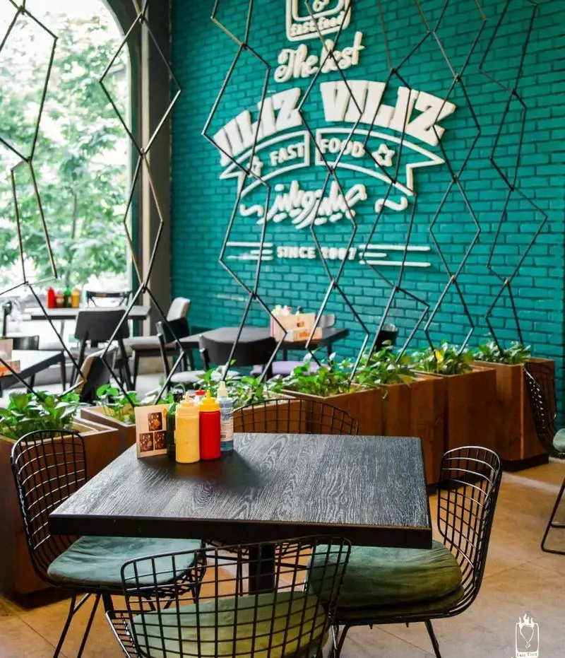 فضای داخلی زیبای فست فود جیلیزویلیز پیروزی با دیوار آجری فیروزه ای و لوگوی سفید، میزهای تیره و صندلی های راحت سبز، همراه با گیاهان سرسبز.