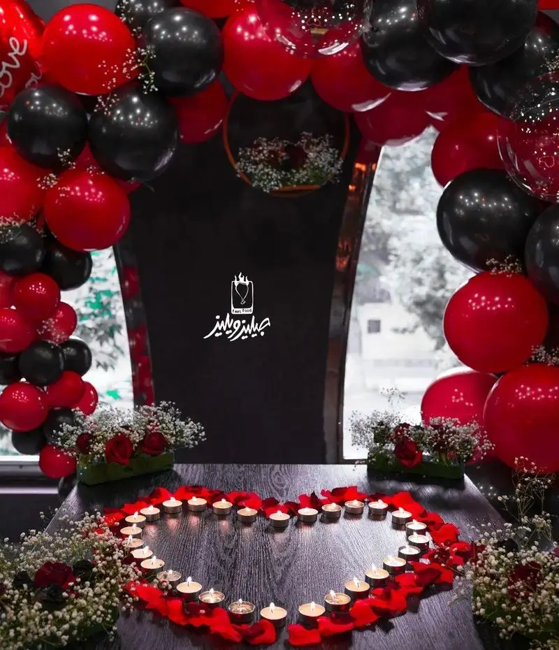 تصویری زیبا از فضای رمانتیک و دلنشین فست فود جیلیزویلیز پیروزی با میز آرایی قلب شمع و بادکنک‌های قرمز و مشکی.