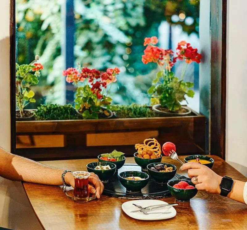 لحظاتی خوش در کافه هاگ: میز صبحانه‌ای دلپذیر با چای داغ، انواع غذاهای تازه و منظره سرسبز گل‌های شمعدانی از پنجره.