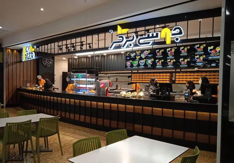 فضای داخلی مدرن و شیک رستوران جوسی برگر با پیشخوان آماده سازی غذا، منوی دیجیتال و میز و صندلی برای مشتریان.