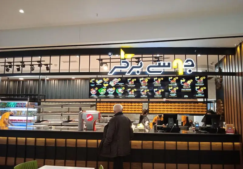 فضای داخلی رستوران جوسی برگر، پیشخوان مدرن با منوهای دیجیتال حاوی انواع برگر و سالاد، مشتری در حال سفارش غذا.