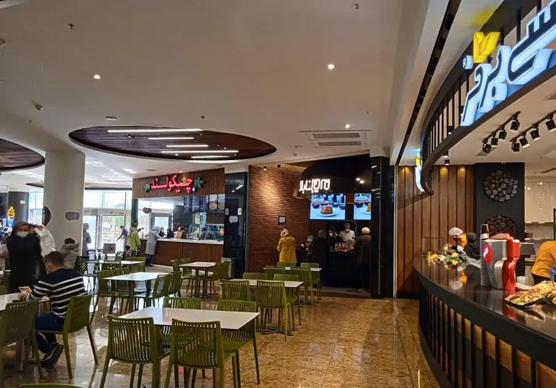 فضای داخلی فودکورت شلوغ و مدرن با تابلوهای روشن، از جمله رستوران جوسی برگر و چیکولند. مشتریان در حال صرف غذا و سفارش.