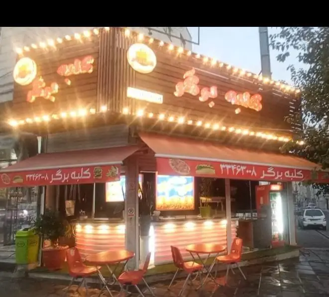 تصویری از نمای شبانه فست فود کلبه برگر با تابلوی روشن، دکوراسیون چوبی و چراغانی جذاب، به همراه میز و صندلی‌های فضای باز نارنجی.