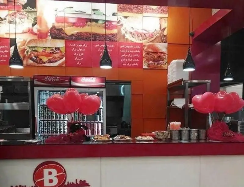 تصویری از فضای داخلی فست فود شهربرگر، شامل منوی همبرگر و ساندویچ، پیشخوان پر از غذا، یخچال‌های کوکاکولا و دکوراسیون جذاب با بادکنک‌های قلبی